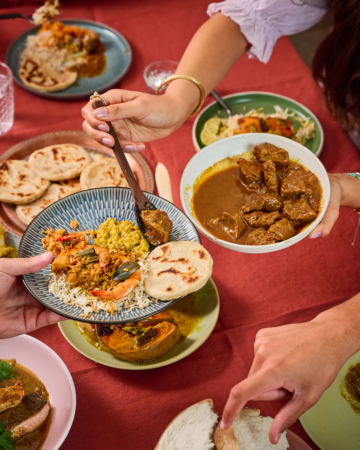 A close-up dining scene showing multiple hands serving and eating a richly spiced meat curry with rice, flatbread, and bread on a red tablecloth.