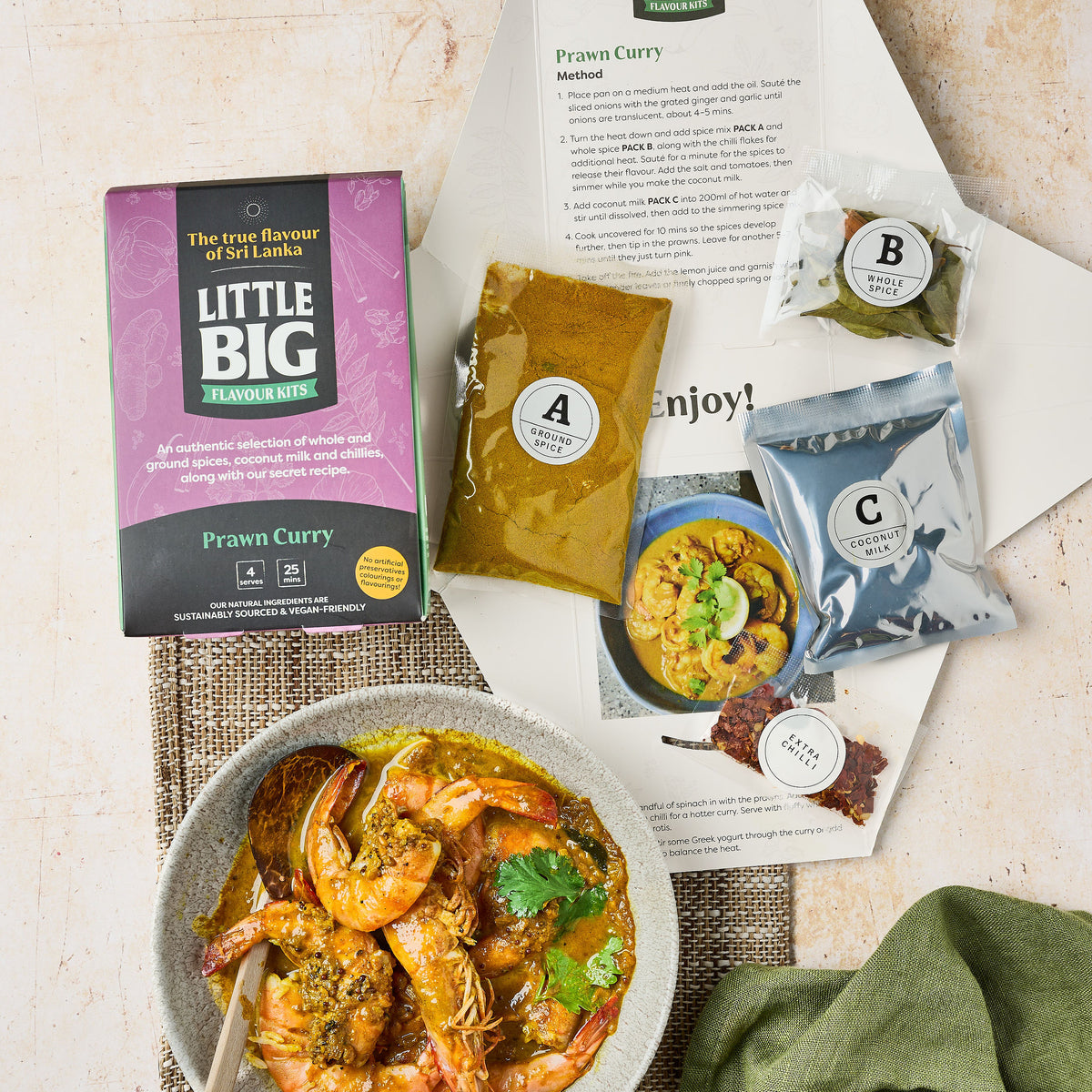 The front of a prawn curry kit. The inside of our prawn curry kit: we're showing the recipe, the A, B, C, and extra chili seasoning packs, as well as a prepared prawn curry on a woven placemat with a green napkin.