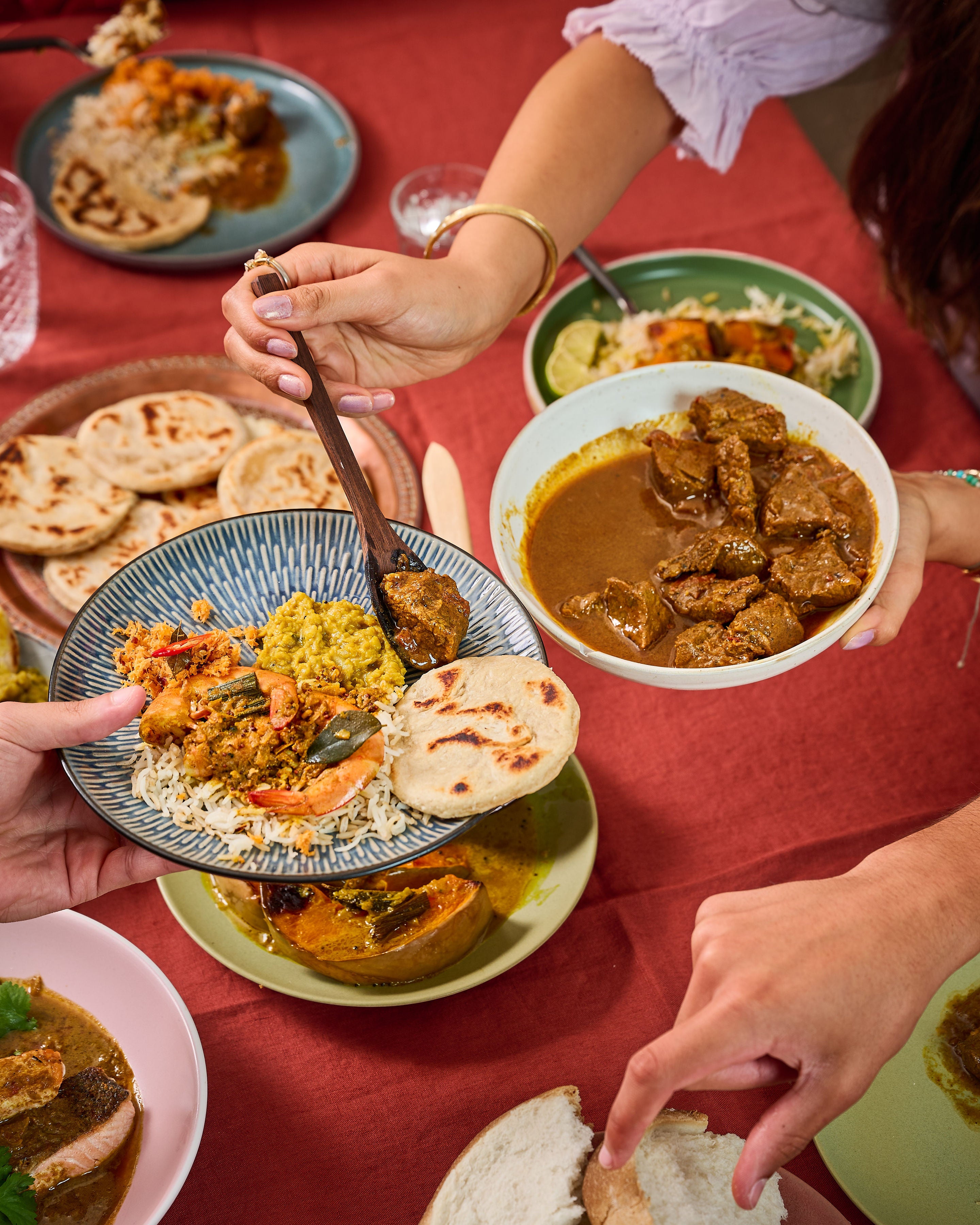 A close-up dining scene showing multiple hands serving and eating a richly spiced meat curry with rice, flatbread, and bread on a red tablecloth.