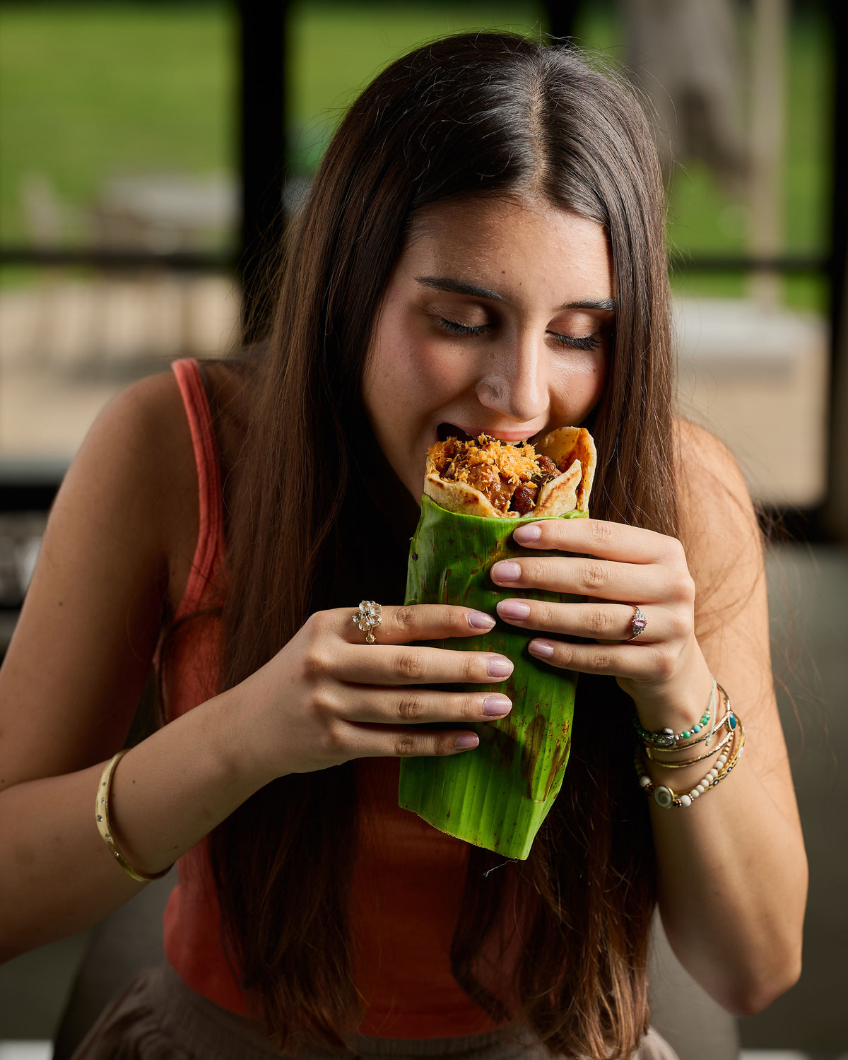 A woman with long dark hair is shown in a close-up shot, eating a wrap filled with meat and vegetables, wrapped in a green banana leaf. She is wearing a rust-orange sleeveless top and holding the food with both hands.