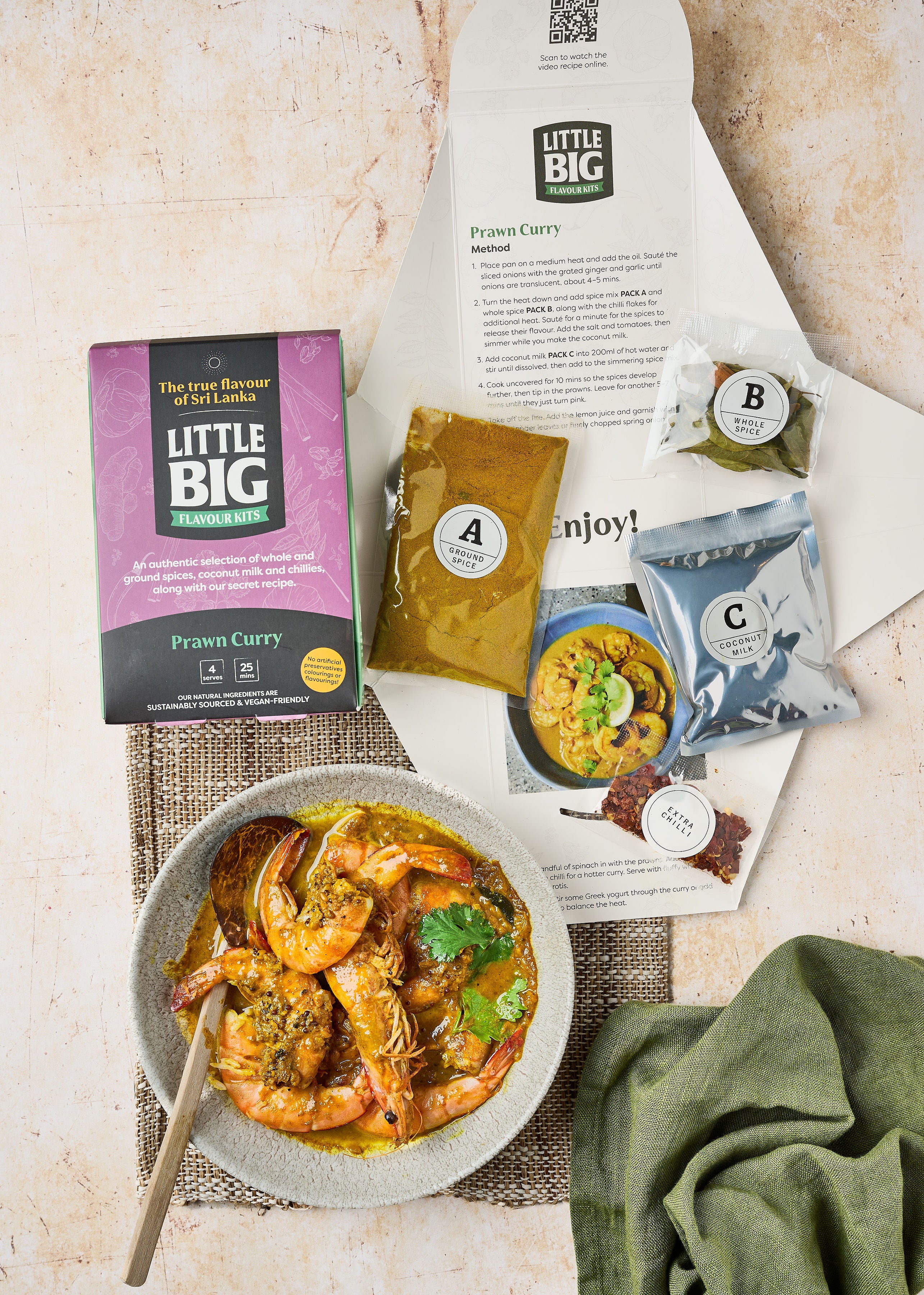 The front of a prawn curry kit. The inside of our prawn curry kit: we're showing the recipe, the A, B, C, and extra chili seasoning packs, as well as a prepared prawn curry on a woven placemat with a green napkin. 