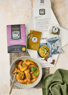 The front of a prawn curry kit. The inside of our prawn curry kit: we're showing the recipe, the A, B, C, and extra chili seasoning packs, as well as a prepared prawn curry on a woven placemat with a green napkin. 