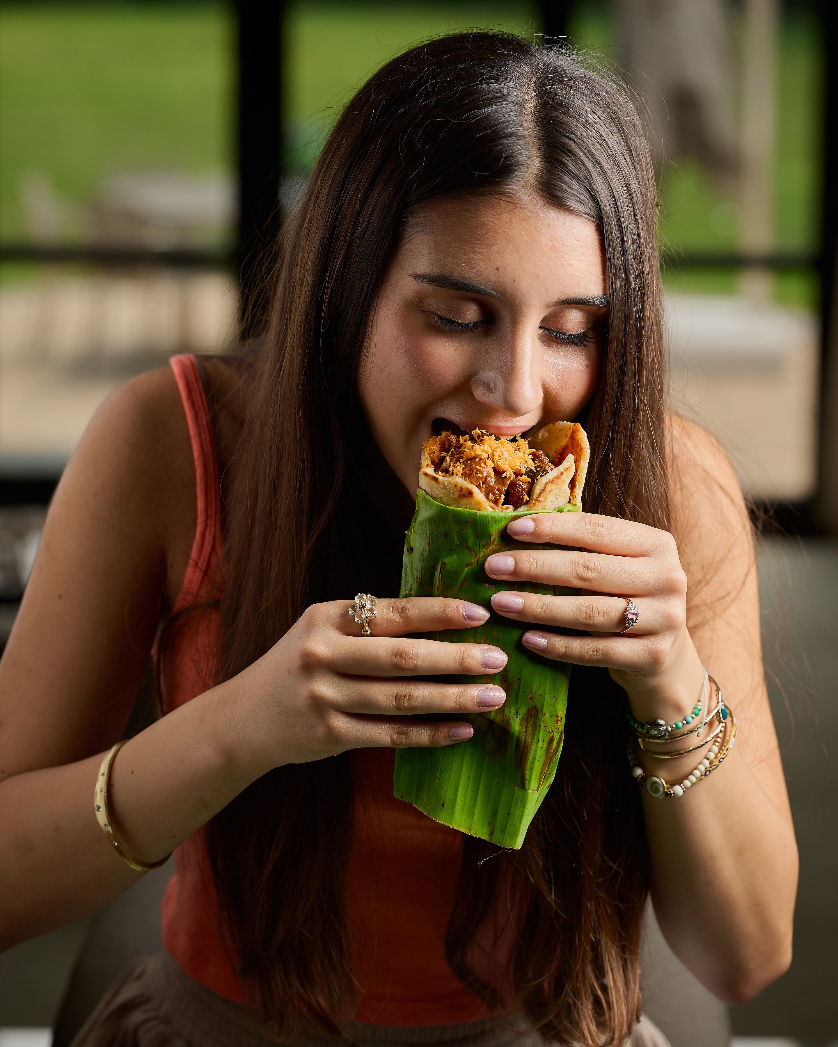 A woman with long dark hair is shown in a close-up shot, eating a wrap filled with meat and vegetables, wrapped in a green banana leaf. She is wearing a rust-orange sleeveless top and holding the food with both hands.