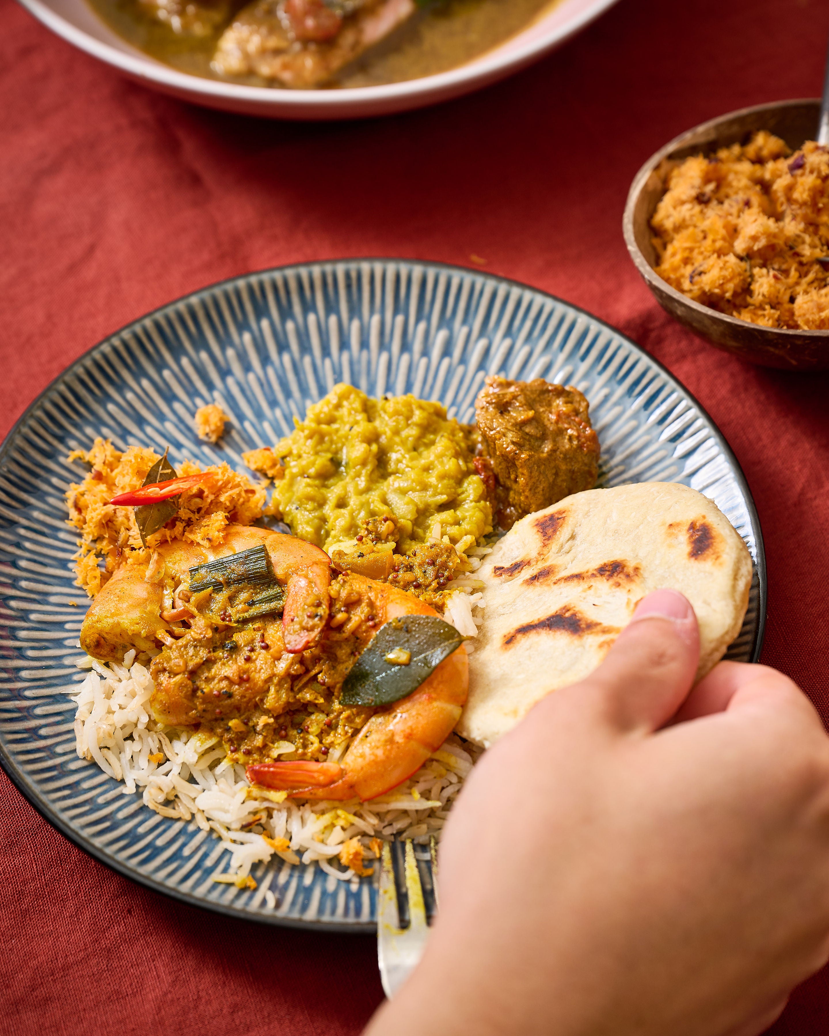 A close-up of a hand holding a piece of flatbread over a blue patterned plate filled with rice, curry, shrimp, and a yellow side dish, set on a red tablecloth with other bowls in the background.